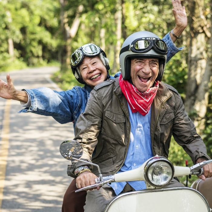 An older couple smiling riding a moped scooter together down a tree-filled street