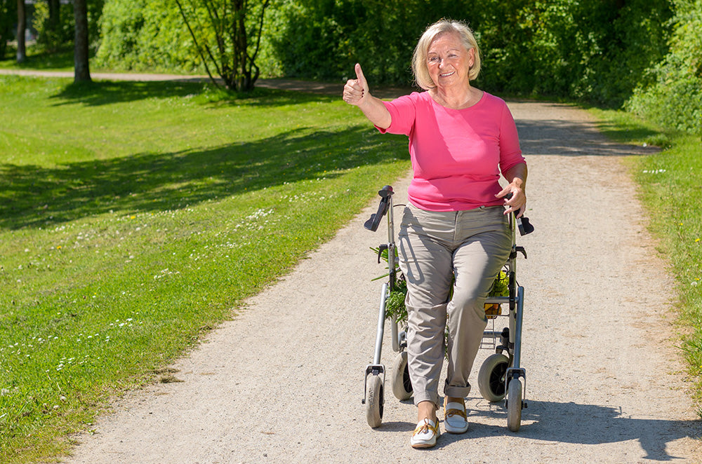 An elderly woman sitting on her walker at a sunny park. She is holding a thumb up and smiling.
