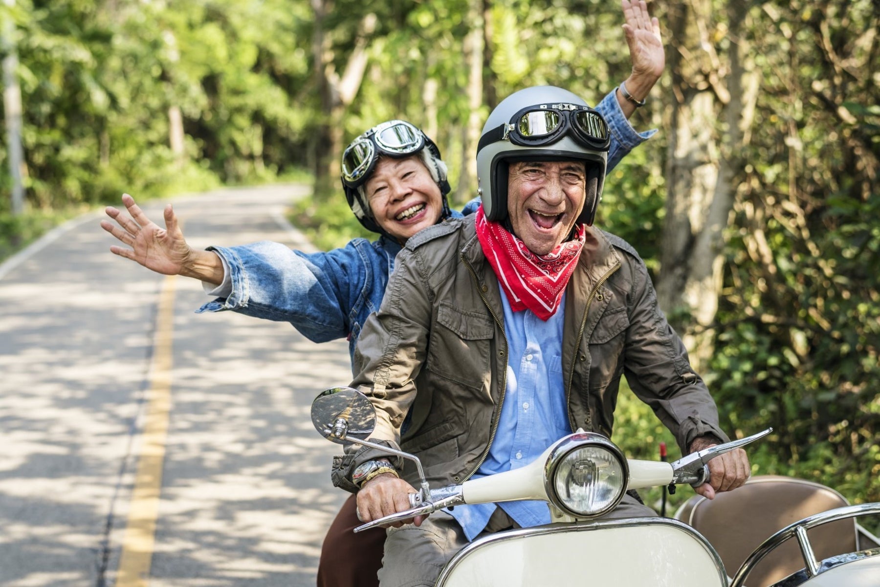 An older couple smiling riding a moped scooter together down a tree-filled street