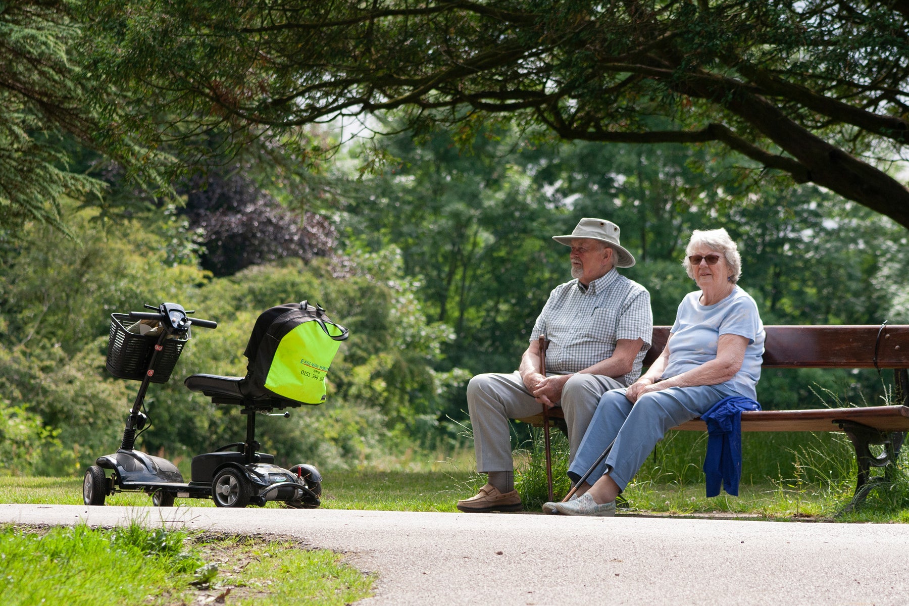 an elderly man and woman sitting on a bench in a park with a mobility scooter next to them