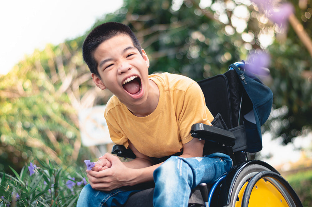 A young boy in a wheelchair smiling outside holding a purple flower 