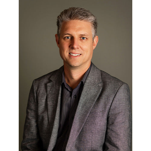 David wearing a grey blazer facing the camera slightly side on and smiling with a dark grey background.