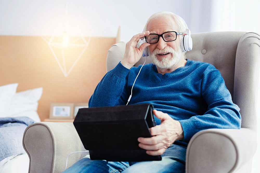 A smiling elderly gentleman sitting on a sofa chair with a tablet in hand and headphones connected
