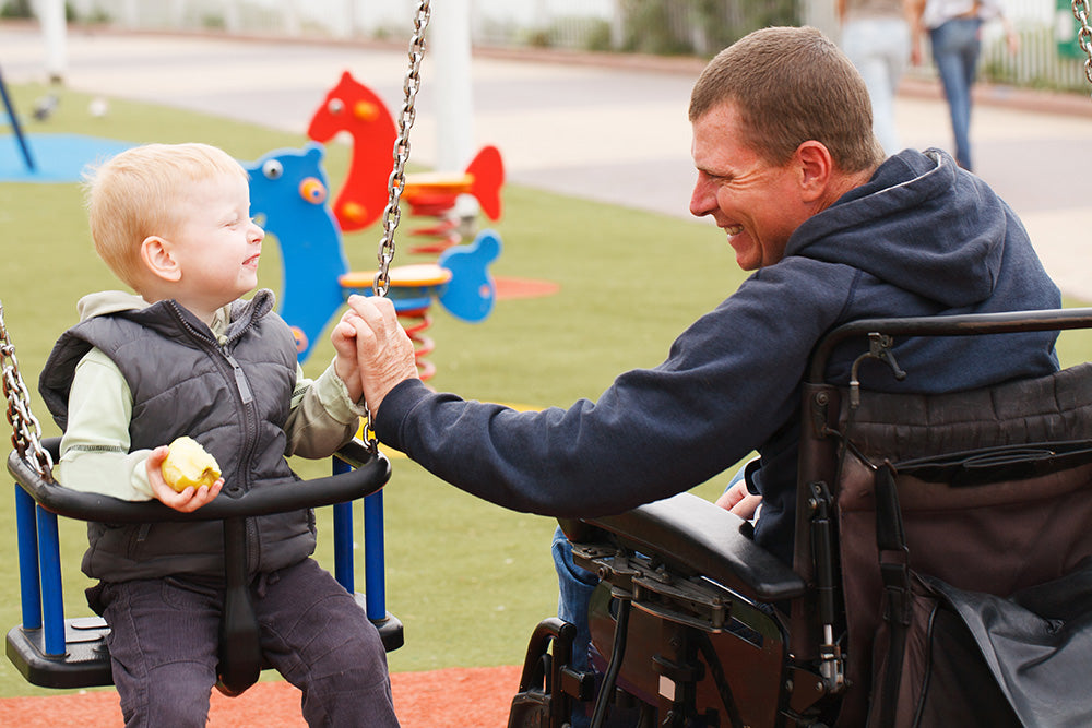 A father in a wheelchair playing with his son on a swing-set at a playground, both are smiling